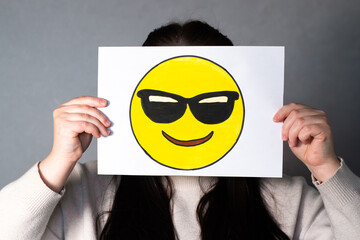 Woman holding happy emoji with sunglasses mask in front of face. Social media trends, Gen Z marketing, World emoji day 17 July.