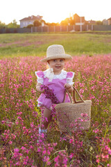 Beautiful one-year-old baby girl stands in a pink dress and a straw hat in a field at sunset among bright flowers.