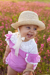Beautiful one-year-old baby girl stands in a pink dress and a straw hat in a field at sunset among bright flowers.
