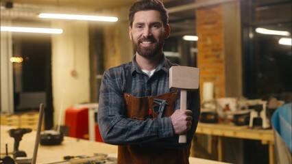 Joyful Caucasian man wood master holding wooden hammer portrait in workshop guy smile male...
