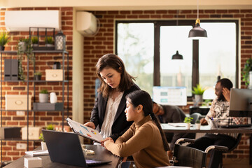 Female coworkers providing insights on business growth, collaborating at office desk with laptop. Caucasian manager with clipboard showing financial graphs to employee in wheelchair, reviewing data.