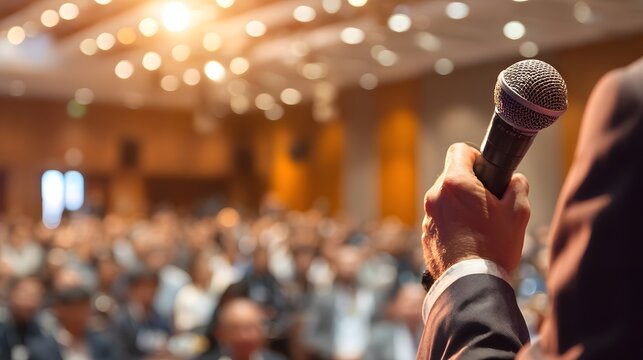 Speaker addressing a large audience, microphone in hand, at a conference or event, communicating information in a professional setting.