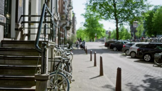 Bicycles resting against railings on a sunlit european city street with defocused background of people walking and parked cars lining the tree-shaded road on a bright day.