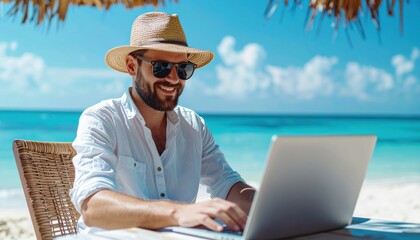 Fototapeta premium Man working remotely on a laptop under a straw parasol at a sunny tropical beach