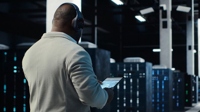 Admin walking in data center, listening music while doing inspection on infrastructure tech. IT specialist in server hub wearing headphones analyzing traffic patterns of equipment mainframes, camera B