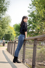 A young teenager girl stands comfortably on a sunlit wooden bridge, casually dressed in a black top, jeans, and black boots.