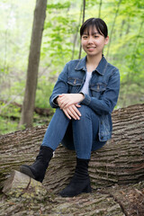 A young teenage girl in denim sits calmly on a fallen tree in a lush green forest, gazing off thoughtfully, surrounded by greenery.