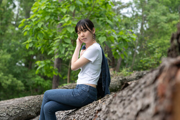 A young teenage girl in a white top and jeans sits calmly on a fallen tree in a lush green forest, gazing off thoughtfully, surrounded by greenery.