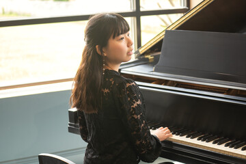 A young teenage girl in a black blouse sits gracefully by a grand piano, smiling at the camera in a bright, elegant room.