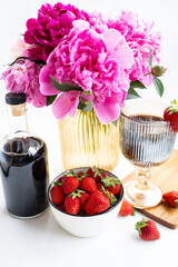A glass of dark wine decorated with strawberries, Pink peony flowers in a vase and a bottle of red wine
