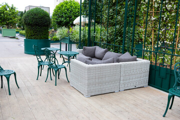 A modern grey sofa with cushions is installed in a glass greenhouse outside. Lush green plants and vibrant flowers surround the living area, creating a serene atmosphere.