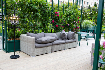 A modern grey sofa with cushions is installed in a glass greenhouse outside. Lush green plants and vibrant flowers surround the living area, creating a serene atmosphere.
