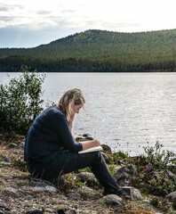 Outdoor woman reading a book while sitting at the shore of a lake on a bright summer day in Sweden. She is enjoying the peace and quiet of nature.