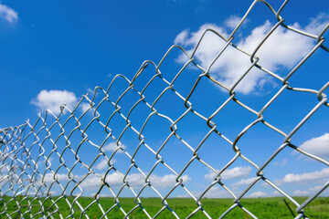 Chain link fence stands against bright blue sky dotted with fluffy white clouds, creating striking contrast. fence metallic texture reflects sunlight, enhancing vibrant outdoor scene