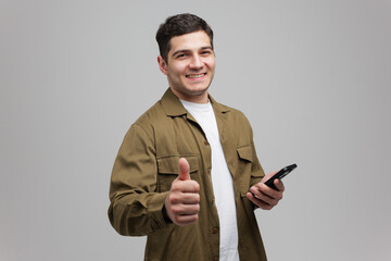 Young man smiling and giving a thumbs up while holding a smartphone in a minimalist studio setting...