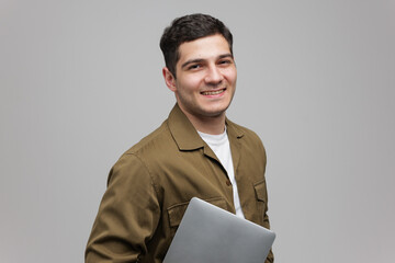 Smiling young man holding a laptop while standing against a light gray background in a modern and...