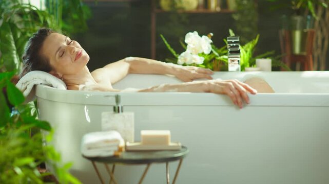 A middle-aged woman unwinds in a tranquil, luxurious bubble bath, surrounded by lush plants. With eyes closed and a serene expression, she embraces a peaceful self-care moment.