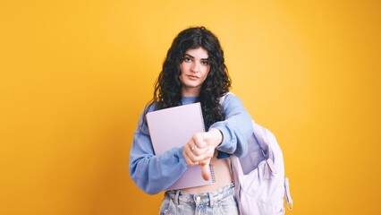 Young student showing thumbs down while holding notebook and backpack on yellow background