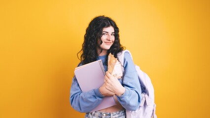 Young student holding money and school supplies on yellow background