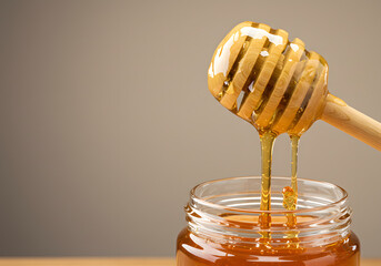 Honey dripping from wooden dipper into glass jar on beige background with golden texture for sweet food and healthy natural product