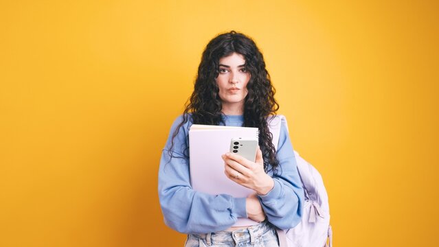 Young student using smartphone and holding books on yellow background