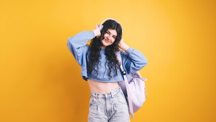 Brunette student listening music with wireless headphones and backpack on yellow background