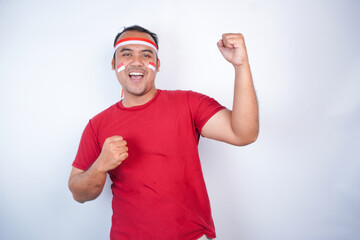Naklejka premium A young Asian man with a happy successful expression, raised a fist wearing red shirt and flag headband isolated by white background. Indonesia's independence day concept.