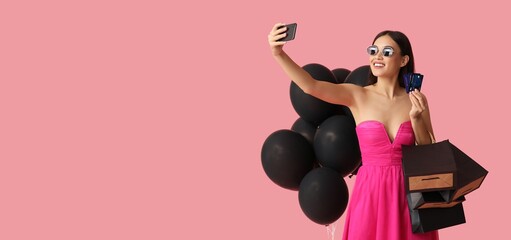 Beautiful young Asian woman with shopping bags, balloons and credit cards taking selfie on pink background with space for text. Black Friday sale