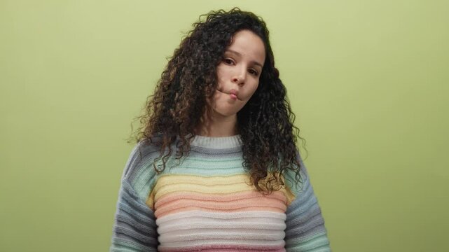 Young hispanic woman with curly hair makes a playful fish face expression against a soft yellow wall background, wearing a colorful sweater, isolated and casual.