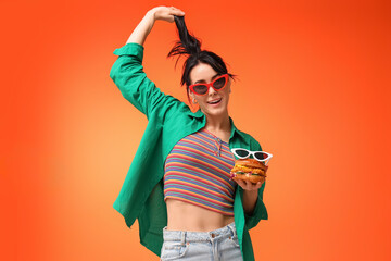 Beautiful young woman with tasty burger and sunglasses on orange background