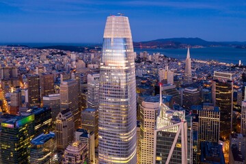 San Francisco, USA, at dusk. The Salesforce Tower dominates the skyline, surrounded by other skyscrapers and city lights. The Golden Gate Bridge is visible in the distance.