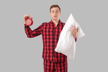 Confused young man with pillow and alarm clock on grey background