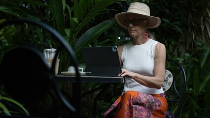 A middle-aged woman sits at a cafe table, focused on her tablet as she works. The lush greenery around her creates a serene atmosphere ideal for a digital nomad.