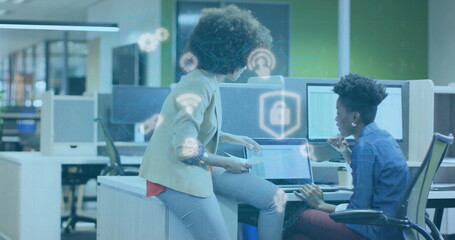 Examining security icons on laptops, two professionals leaning over desk in modern open-plan office