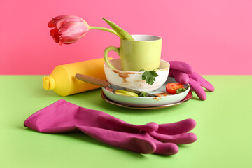 Many dirty dishes with rubber gloves, detergent and tulip flower on color background