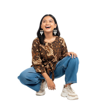 A young woman with a batik top and jeans squatting and looking up with a happy expression on her face