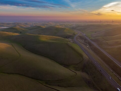 Highway 580 cuts through rolling hills dotted with wind turbines at sunset in The Altamont Pass, Livermore, California, USA. Cars and trucks travel along the highway.