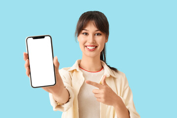 Smiling young woman pointing at blank mobile phone on blue background