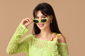 Stylish young woman in green crochet top with sunglasses on beige background, closeup
