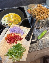 Fresh vegetables, sliced chili, garlic, and shallots prepared on a cutting board, with tofu cubes...