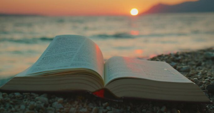Open book Holy Bible on a pebble beach at sunset with sea waves in background