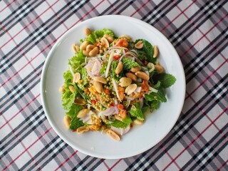 Thai-style Horseshoe Crab Eggs Spicy Salad on a white plate, garnished with fresh herbs, peanuts, and vegetables. The dish is placed on a checkered tablecloth, showcasing vibrant colors and textures.