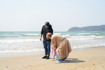 In early March, a 1-year-old Japanese girl walks on a sandy beach as her parents in their 30s hold her hands. Together they enjoy a joyful spring moment by the sea. A heartwarming family scene.