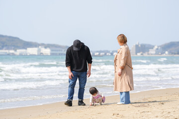 In early March, a 1-year-old Japanese girl explores the beach with curiosity while her parents in their 30s watch her closely. A warm springtime family moment by the sea filled with connection.