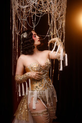 Elegant young circus actress in golden corset and gloves, standing under chandelier, front view....