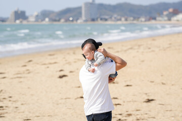 In early March, a Japanese father in his 30s carries his 6-month-old baby on his shoulders while walking along a sandy beach by the sea. A warm springtime scene of family, joy, and connection.