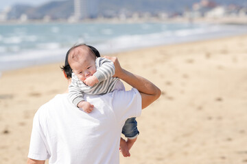 In early March, a Japanese father in his 30s carries his 6-month-old baby on his shoulders while walking along a sandy beach by the sea. A warm springtime scene of family, joy, and connection.