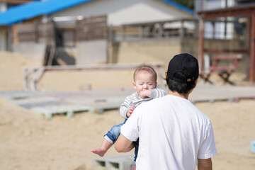 In early March, a Japanese father in his 30s carries his 6-month-old baby on his shoulders while walking along a sandy beach by the sea. A warm springtime scene of family, joy, and connection.