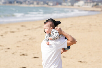 In early March, a Japanese father in his 30s carries his 6-month-old baby on his shoulders while walking along a sandy beach by the sea. A warm springtime scene of family, joy, and connection.