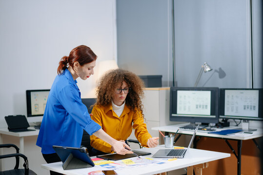 Two women discussing mobile app wireframes in a modern office. UX UI team collaboration, design thinking, and creative planning.